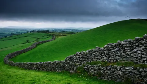 A stone wall curves through lush green hills under a stormy sky with a wind turbine in the distance.