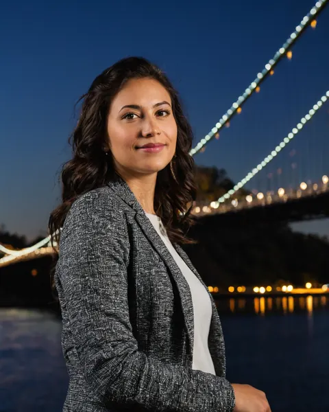 A woman in a gray blazer stands by a lit bridge at twilight over water.