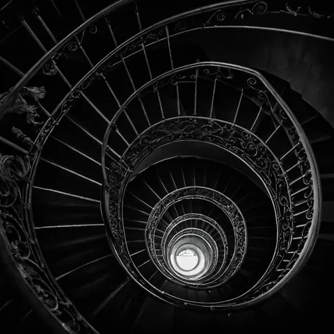Black and white spiral staircase with ornate iron railings leading downward to a bright circular opening.