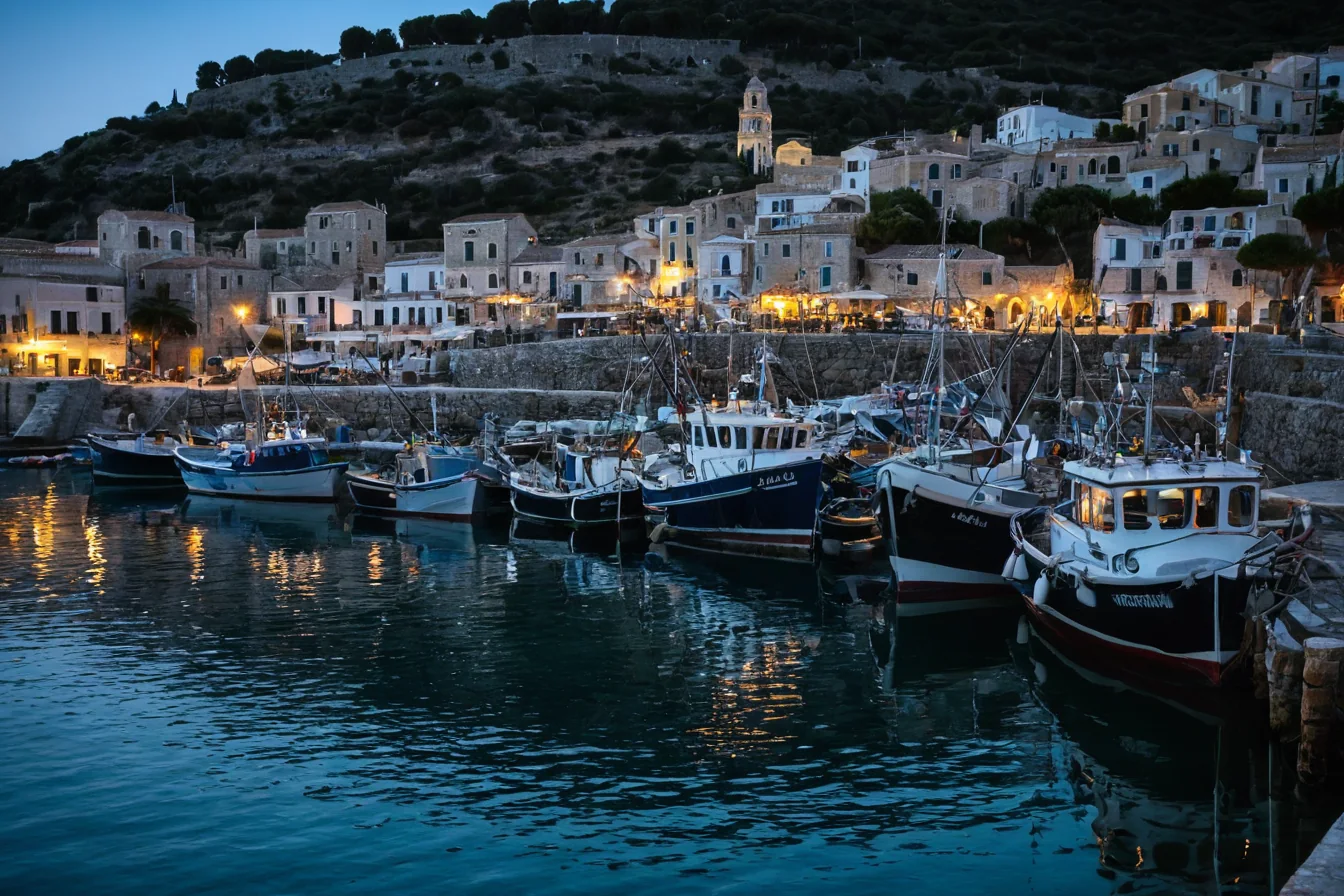 Fishing boats docked in a harbor with glowing lights from coastal buildings at dusk