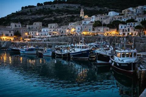 Fishing boats docked in a harbor with glowing lights from coastal buildings at dusk