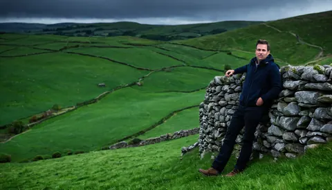 Man leaning on stone wall in green countryside under cloudy sky
