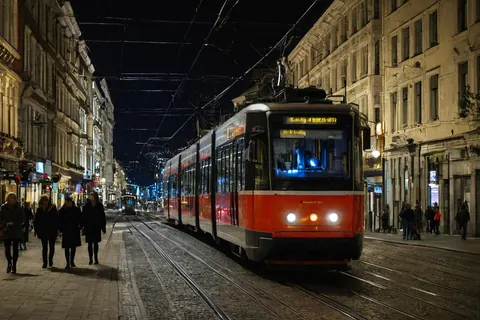 Red tram moving through a historic European city street at night with pedestrians and warm building lights.