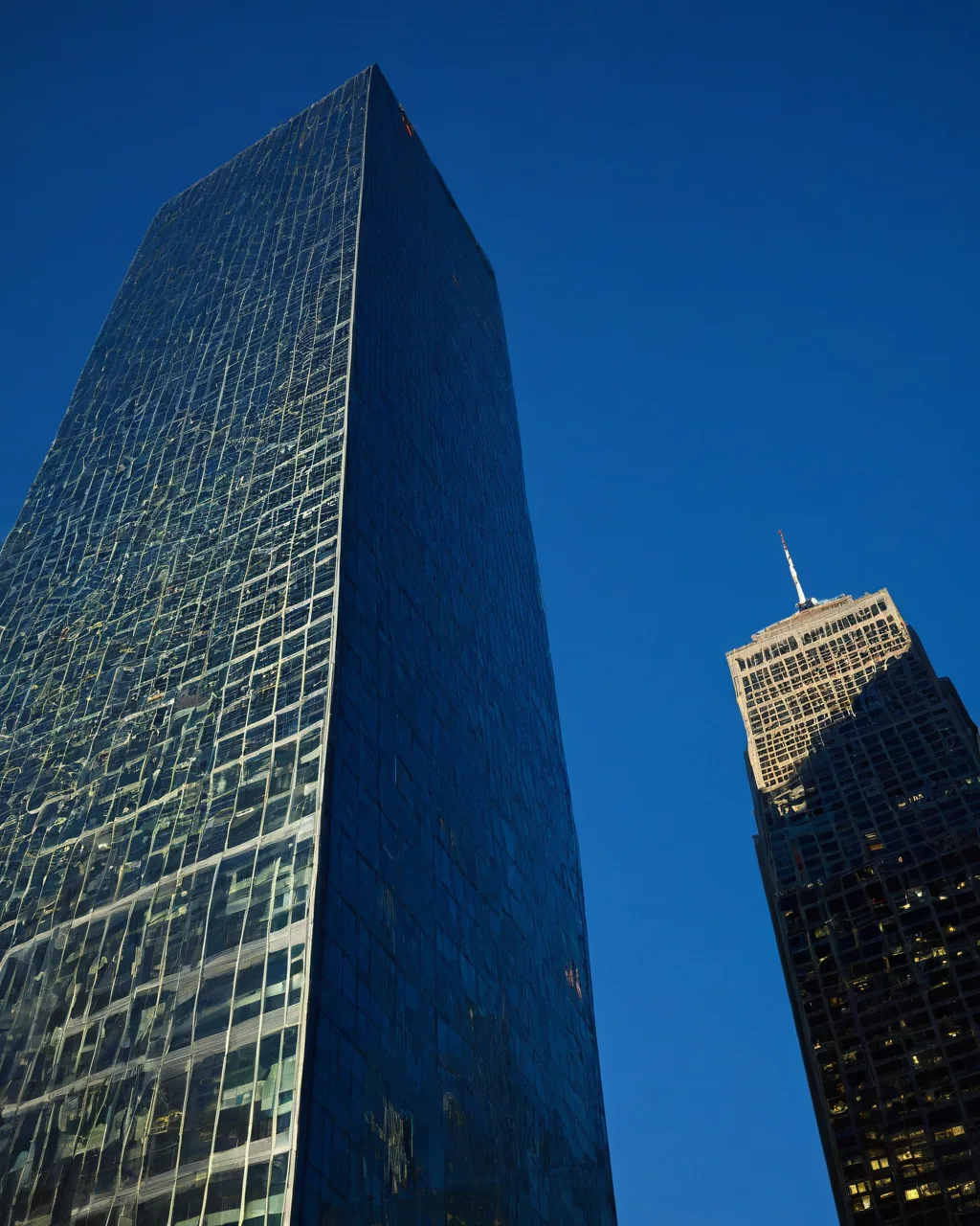 Two tall skyscrapers rise against a clear blue sky one with reflective glass the other with lit windows and antenna