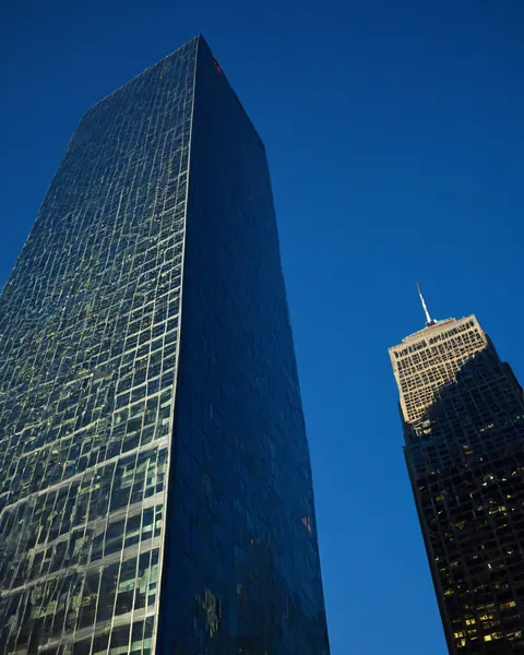 Two tall skyscrapers rise against a clear blue sky one with reflective glass the other with lit windows and antenna