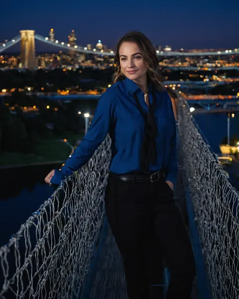 Woman in blue shirt and black pants leaning on bridge railing at night with city lights behind her.